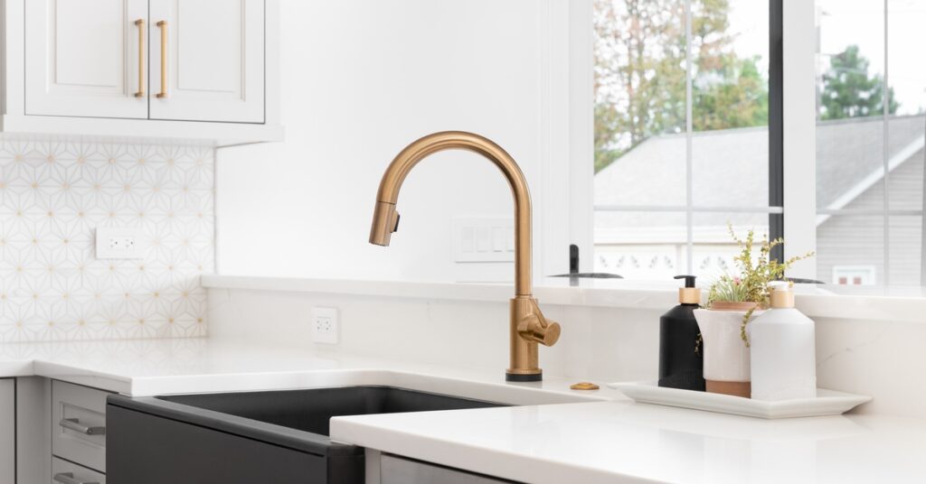 A black farmhouse sink with a copper faucet in a kitchen with white granite countertops and grey cabinetry.