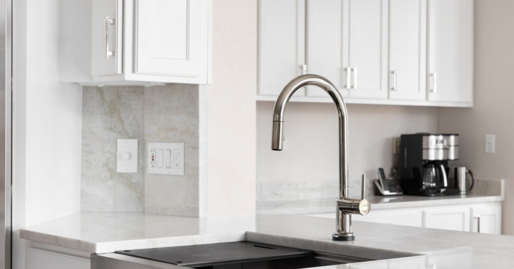 A stainless steel apron-front sink with a stainless steel faucet in a kitchen with marble countertops and white cabinetry.
