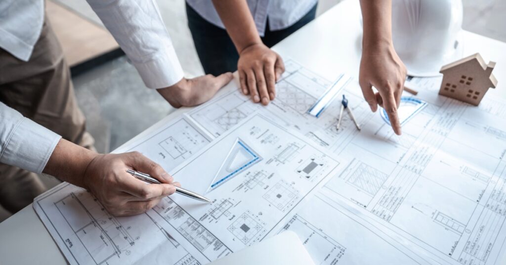 A close-up of a table full of blueprints and home construction plans as two people point at the documents.