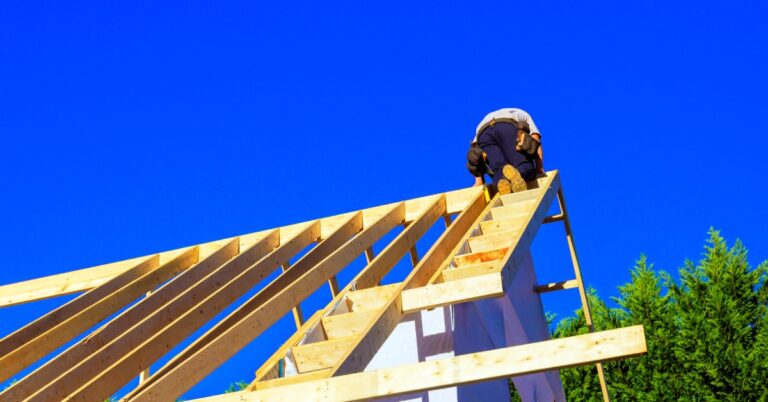 A ground-level view looking up at a worker on the framework joists of a home's unfinished roof against a clear blue sky.