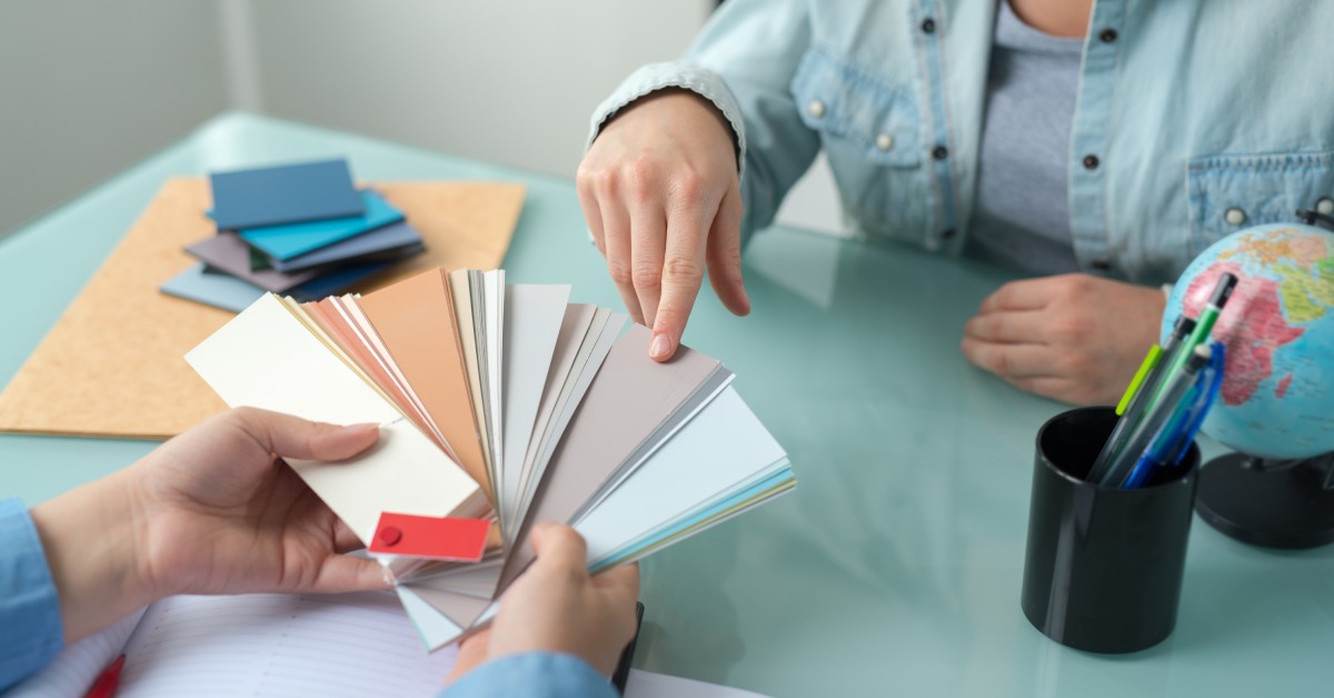 A close-up of two people sitting at a colored table, with one showing the other a bundle of color swatches.