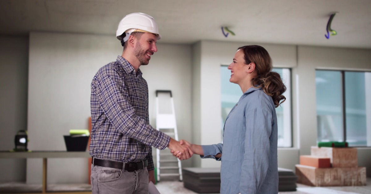 A man in a flannel shirt and a hard hat smiles while shaking the hands of a smiling woman in a home under construction.