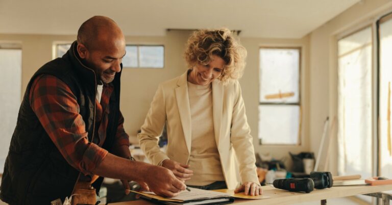 A woman in a white blazer is looking at papers on a workbench inside a home under renovation with a contractor.