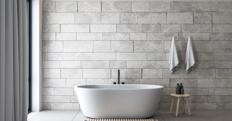 A modern bathroom with a white ceramic bathrumb, gray wall tiling, and hooks with white towels next to a window.