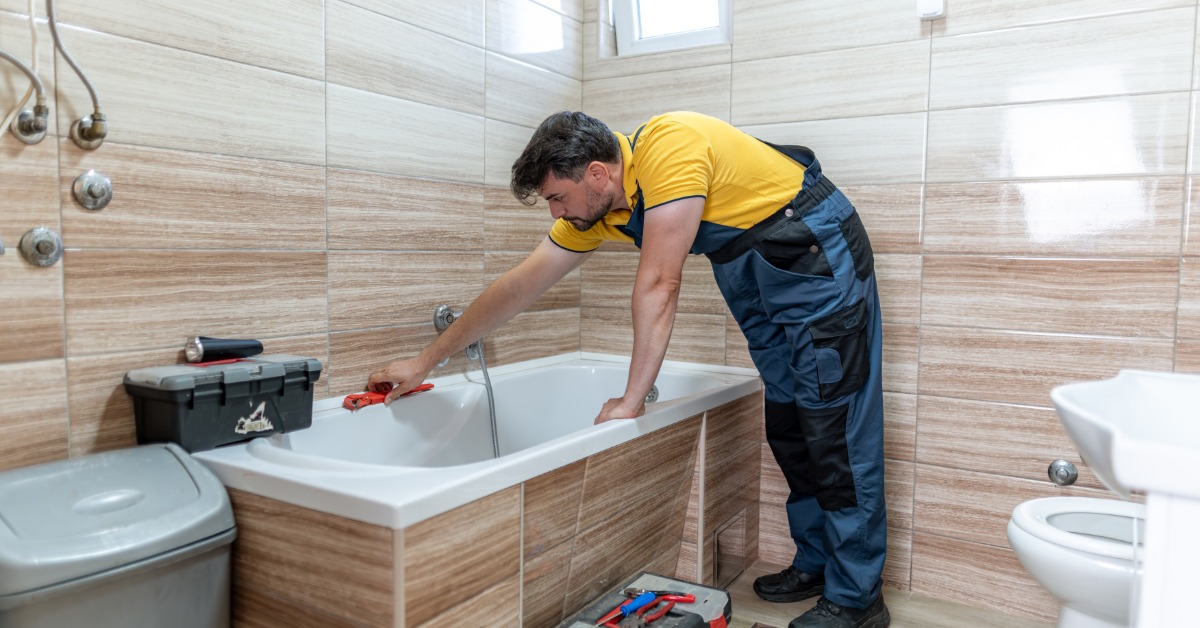 A man in blue overalls and a yellow shirt working on the bathtub in a contemporary bathroom with hand tools.