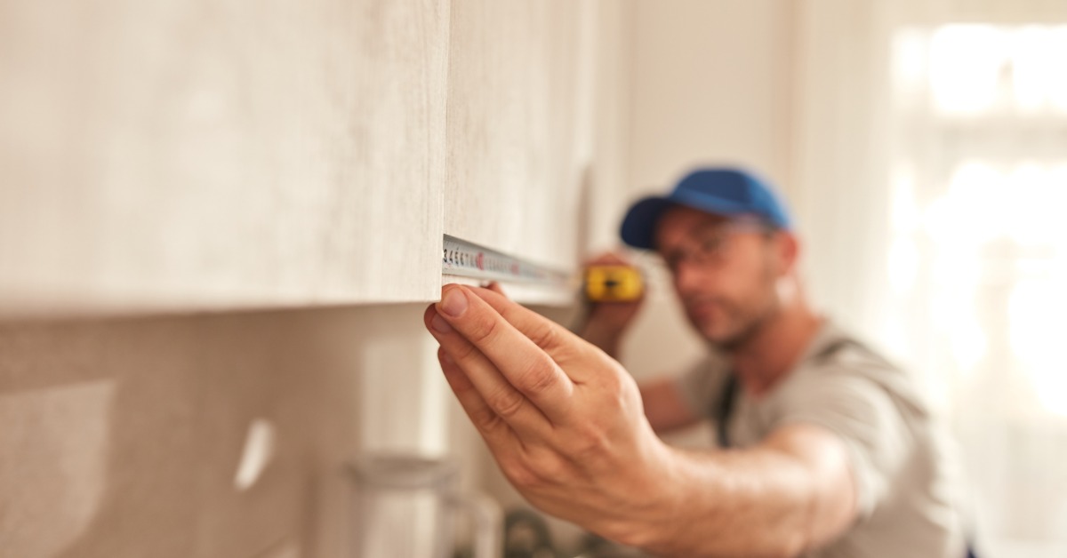 A close-up of a man in a grey shirt and blue hat using a tape measure to measure the upper cabinets of a kitchen.