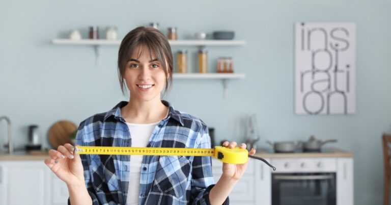 A woman in a flannel shirt with brown hair stands in a home kitchen and holds out a tape measure, partially unraveled.