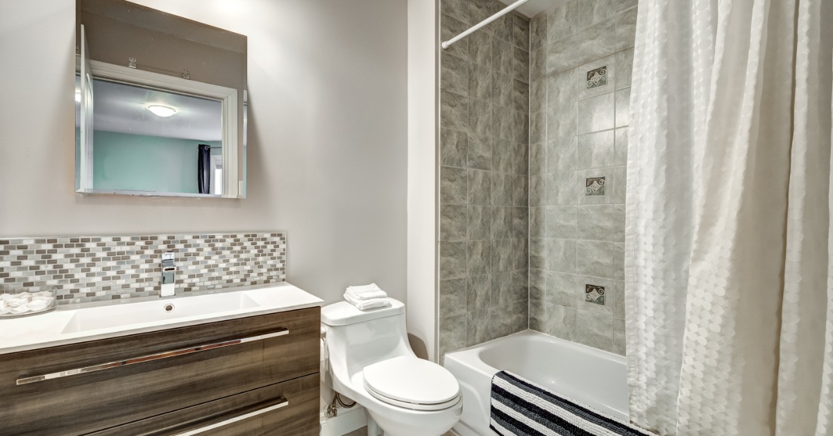 A modern home bathroom with a sink with modern cabinets, tile backsplash, a mirror, and a bathtub/shower next to the toilet.