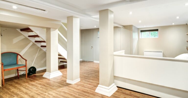 A finished residential basement with hardwood floors, white structural pillars, and a floating staircase leading upstairs.