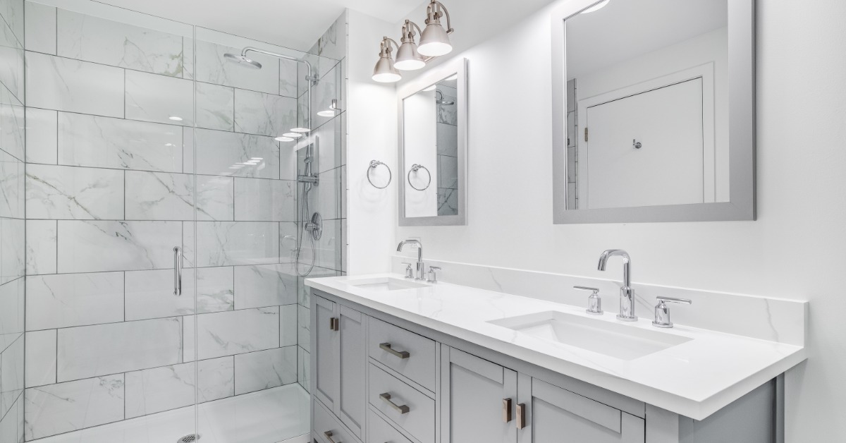 A modern bathroom with two vanity sinks, white marble countertops, grey cabinets, and a shower with a glass door.
