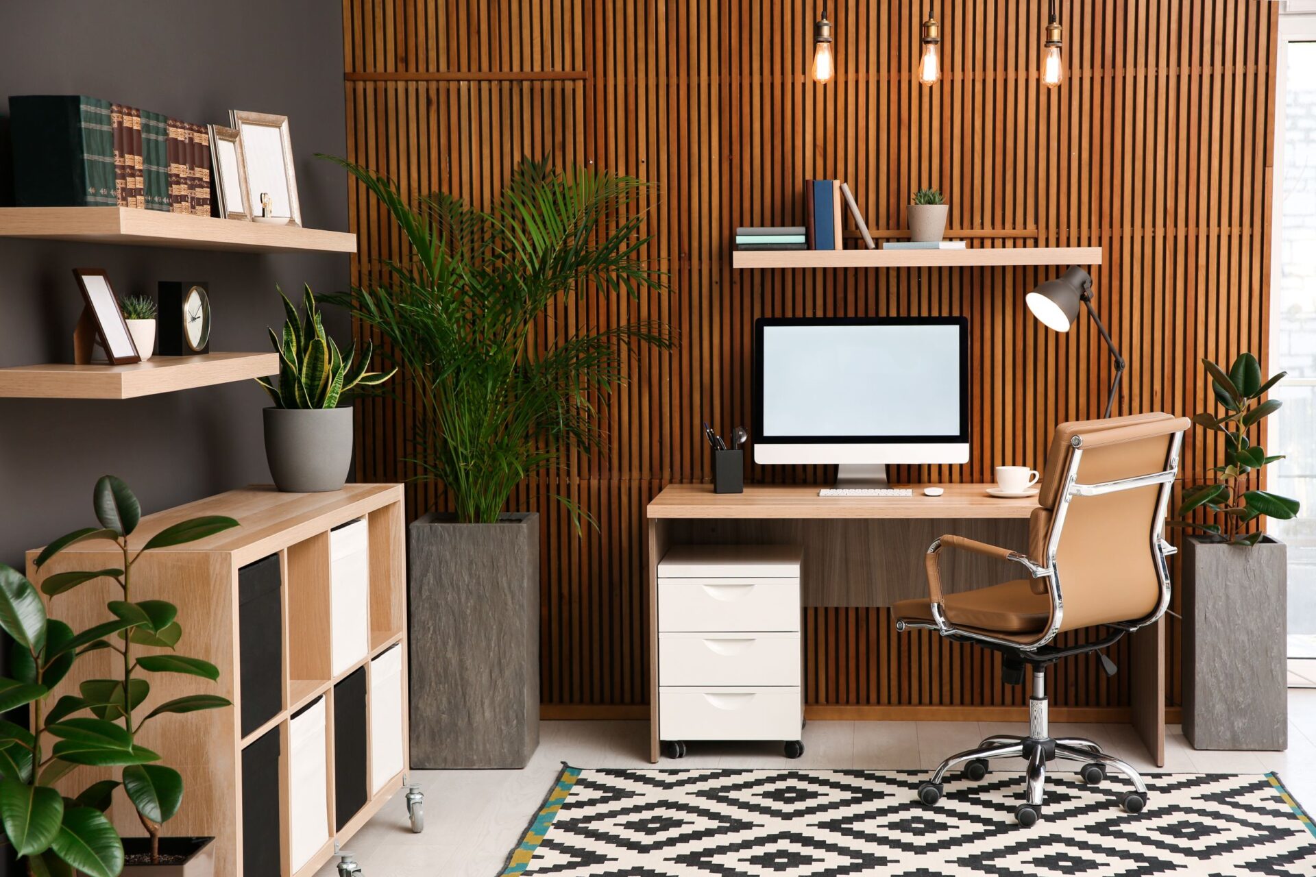 A modern home office with a wooden desk, black chair, floating shelves, planter, bookcase, and wastebasket.