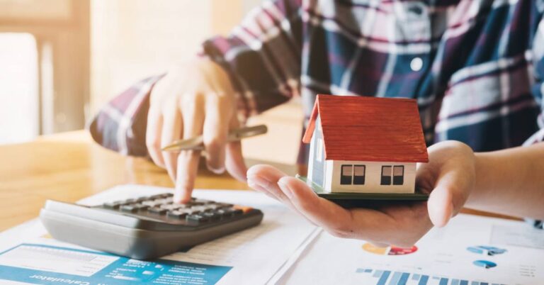 A close-up of a woman sitting at a desk holding a model miniature home in one hand and using a calculator with the other.