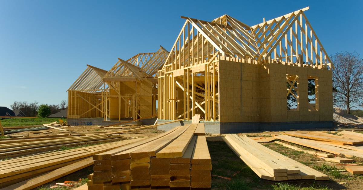 A ground-level view of piles of lumber for home construction on grass in front of a home under construction.