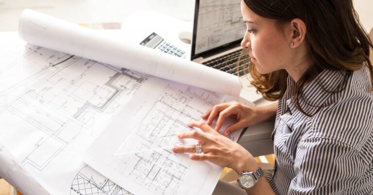 A young adult woman sitting at a desk with numerous blueprints and designs for a home next to a laptop.