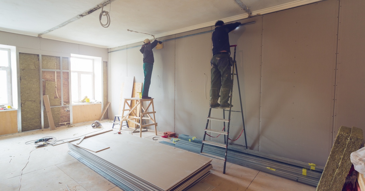 Two contractors on ladders in a house under renovation installing drywall on the wall during the day.