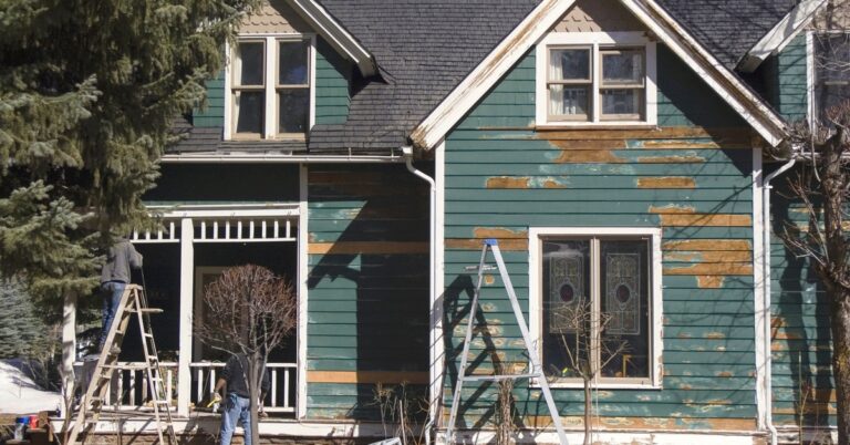 A view from the street of the front of an old green house with chipping paint, with ladders, supplies, and contractors.