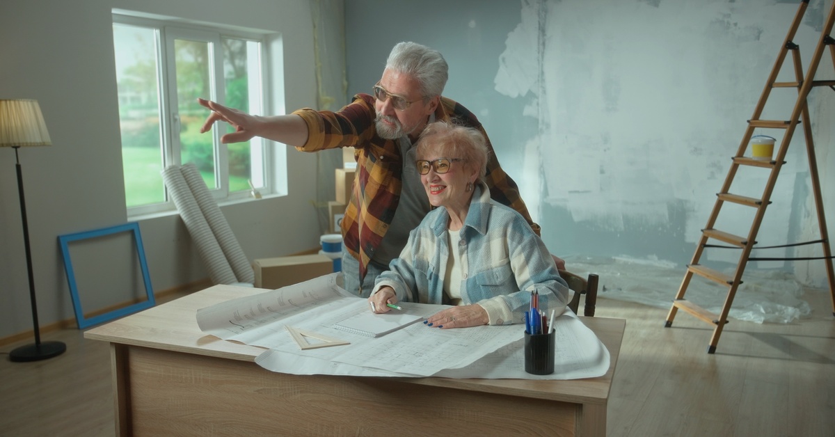 A senior woman sits at a desk in the middle of a room undergoing renovation, as an older man points to something.