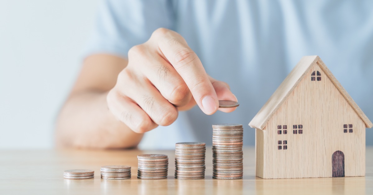 A close-up of a man in a blue shirt placing coins onto a series of coin stacks next to a miniature house model.