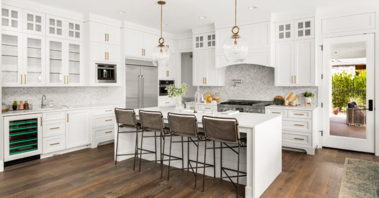 A luxurious home kitchen with white cabinetry, dark hardwood floors, and a kitchen island with quartz waterfall counters.