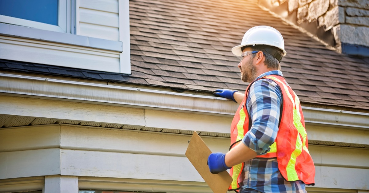 A man in jeans, a flannel shirt, safety glasses, and a hard hat touches the shingles of a roof while holding a clipboard.