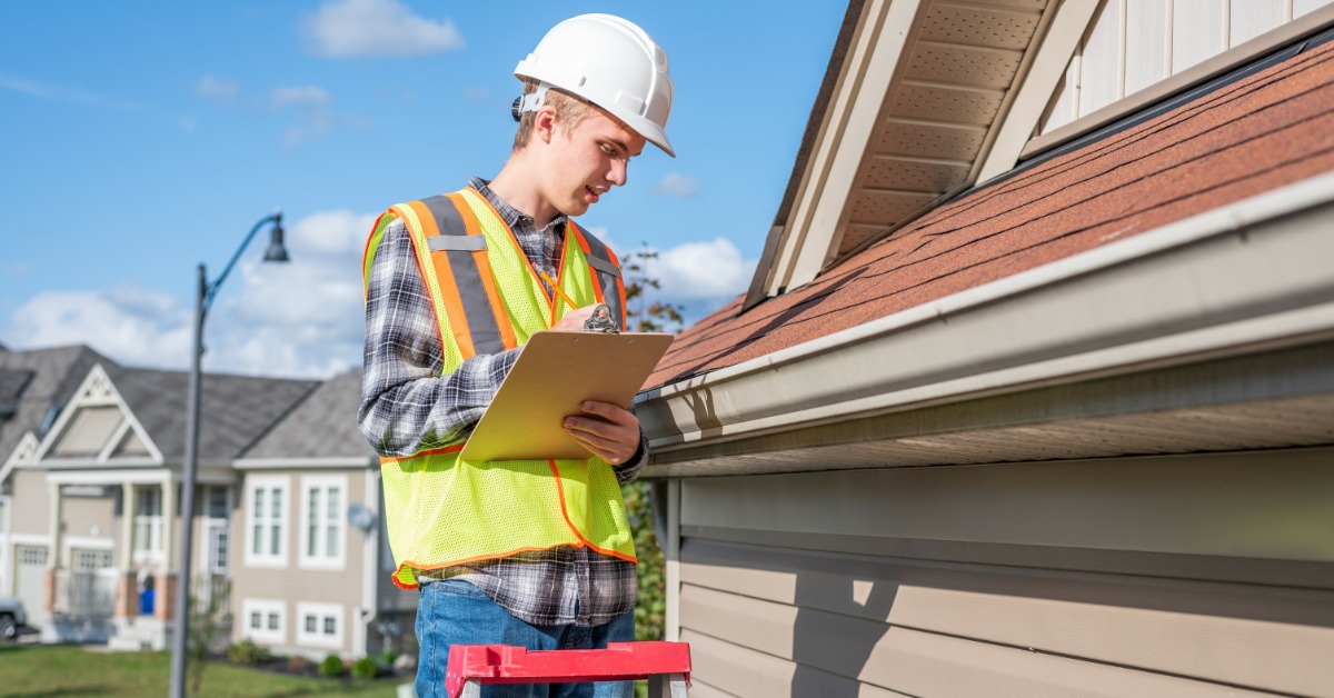 A man in a flannel shirt with a high-visibility vest, hard hat, and clipboard stands on a ladder and examines a home roof.