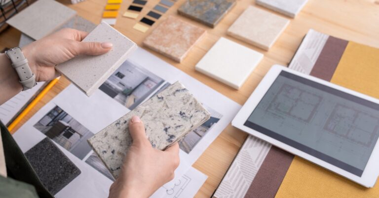 An over-the-shoulder view of a woman holding two samples of stone tiles over a desk with more samples and photographs.