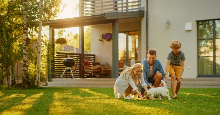 A man, a woman, and a young boy are playing in the backyard grass of a home with a white and brown puppy dog.