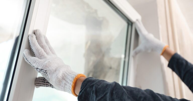 A close-up of a person with white gloves holding up a new window for installation into an indoor frame.