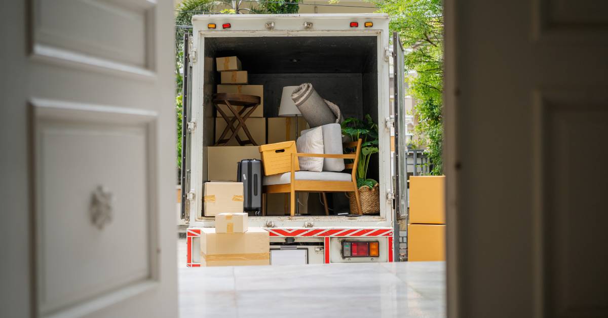 A view looking out from the front door of a home toward a moving truck with the door open, with furniture and boxes inside.