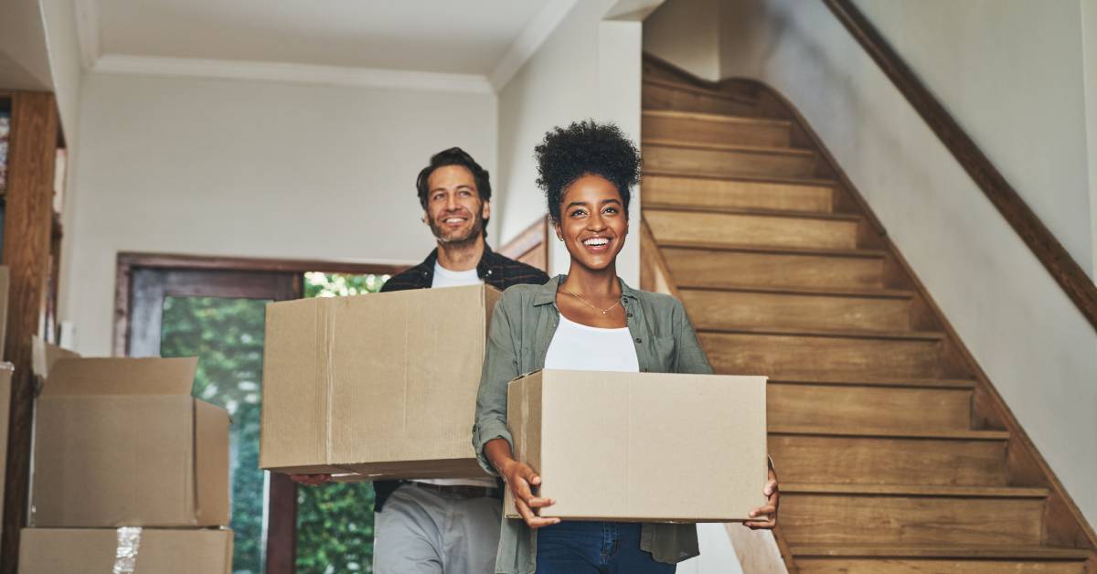A man and a woman walking into a house, smiling while holding cardboard boxes. Next to them are more boxes and a staircase.