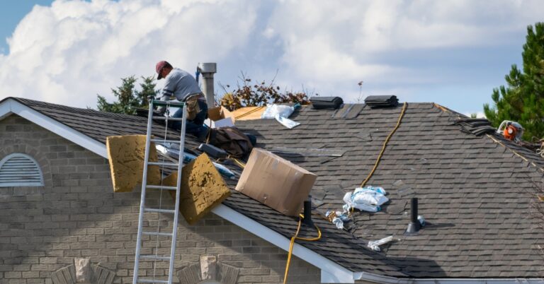A long-distance view of a contractor on the roof of a home on a sunny day with various tools and equipment around him.