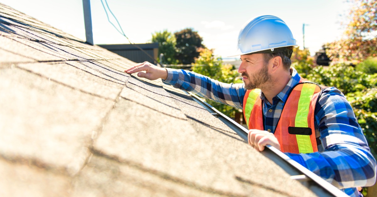 A side view of a man in a flannel shirt, high-visibility vest, and hard hat inspecting the shingles of a roof.