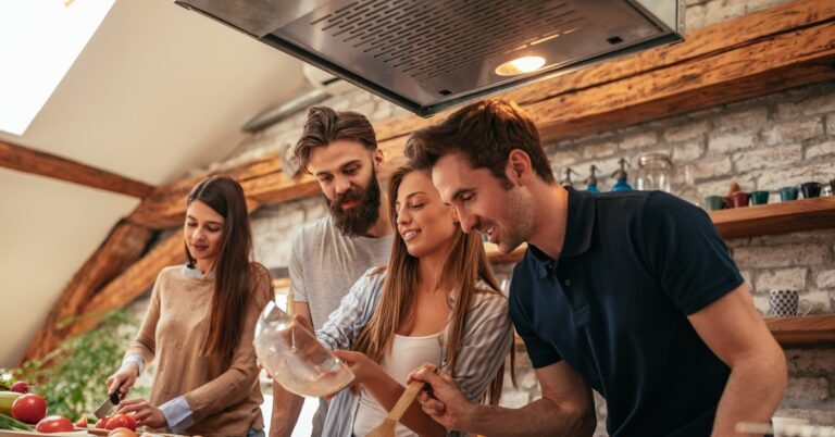 A group of four young people, two men and two women, cooking together in a home kitchen under a vent hood.