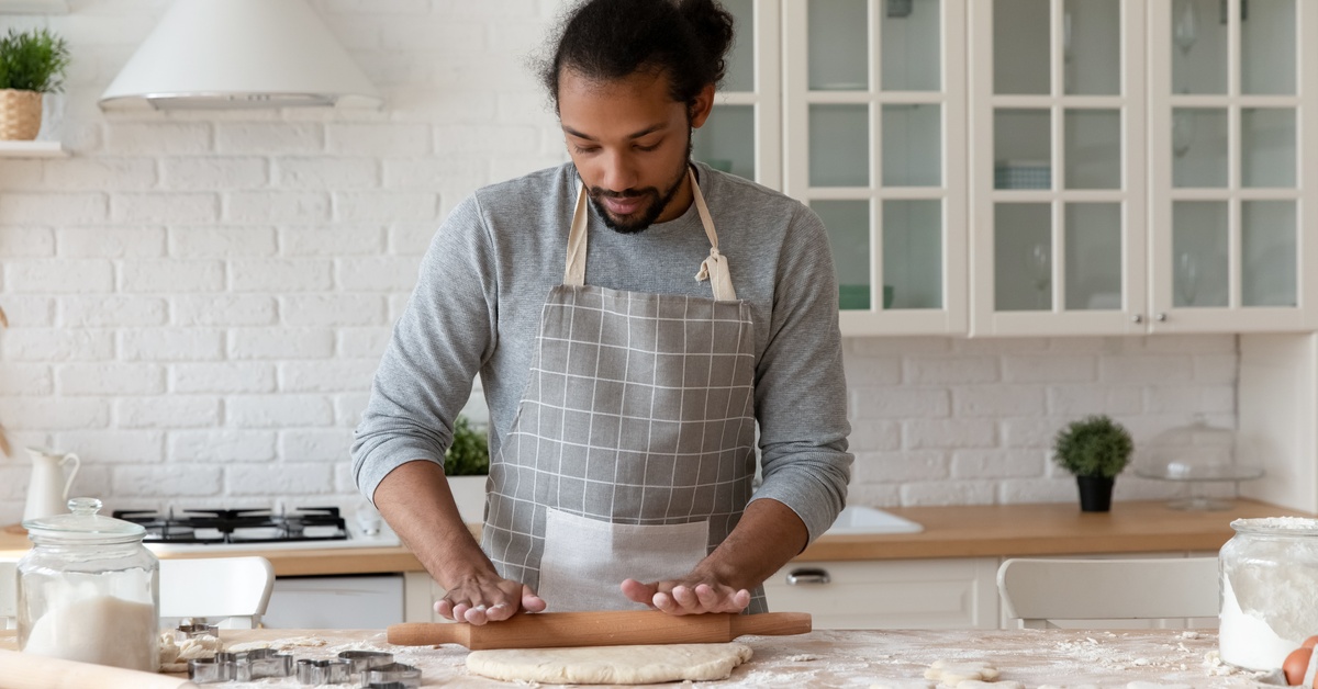 A man wearing a gray apron in a white kitchen using a roller to press dough on the counter of a kitchen island.
