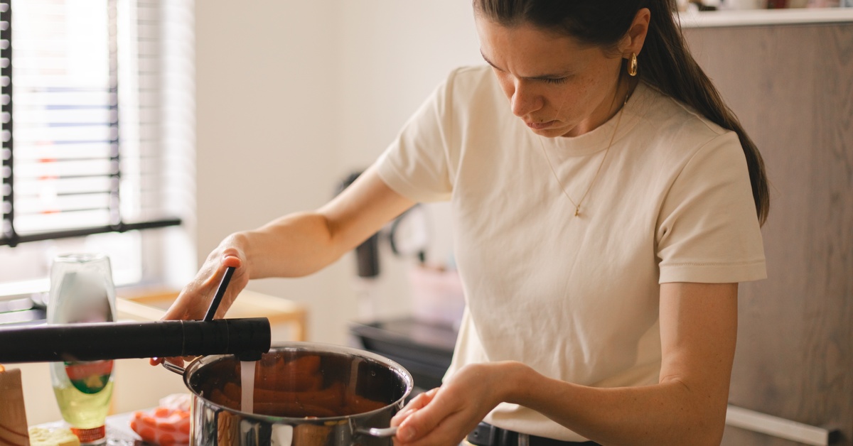 A woman in a white T-shirt and jeans holds a stainless steel pot under a faucet in the kitchen to fill it with water.