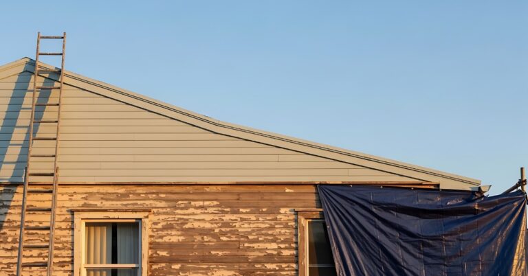 A view from the side of a house with peeling brown paint and a blue tarp draped over a window and a ladder to the roof.