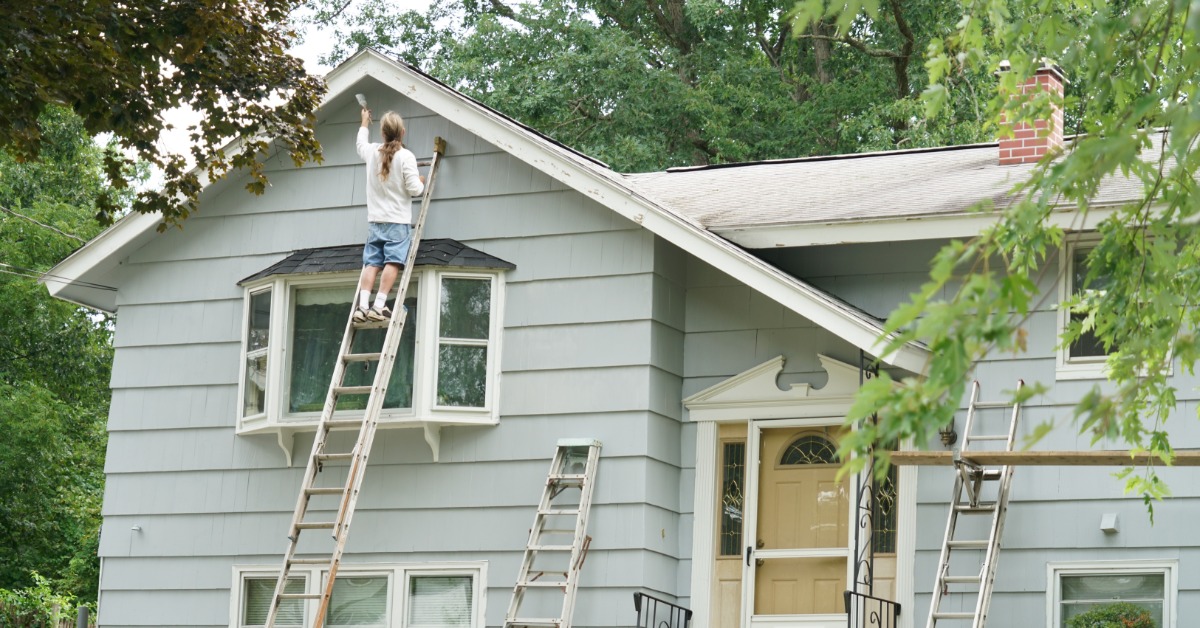 A man standing on a ladder against the side of a house, painting the exterior and roof with a paintbrush.