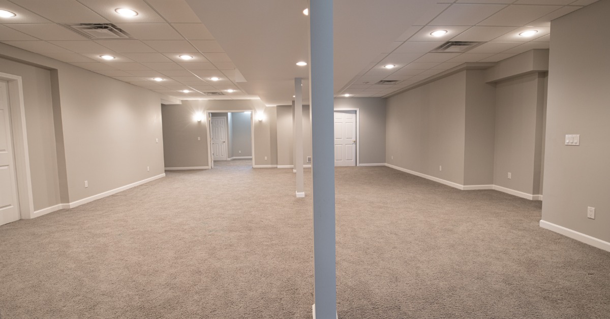 A full view of a finished basement with grey carpet, grey walls, white doors to various rooms, and acoustic ceiling tiles.