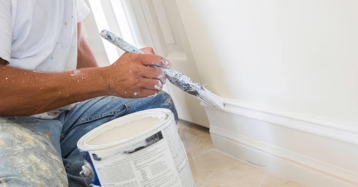 A close-up of a man sitting on a tile floor in paint-splattered jeans and using a white paintbrush to paint baseboard trim.