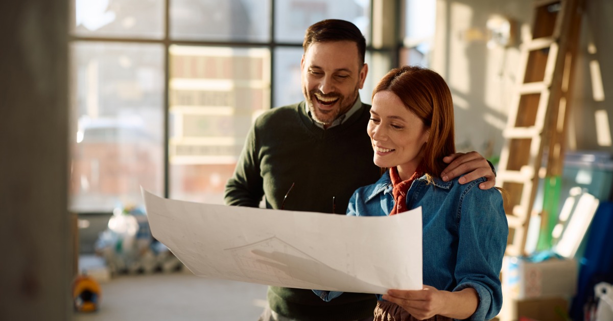 A man and a woman, with the man holding his arm around the woman's shoulders, smile while looking at house plans.
