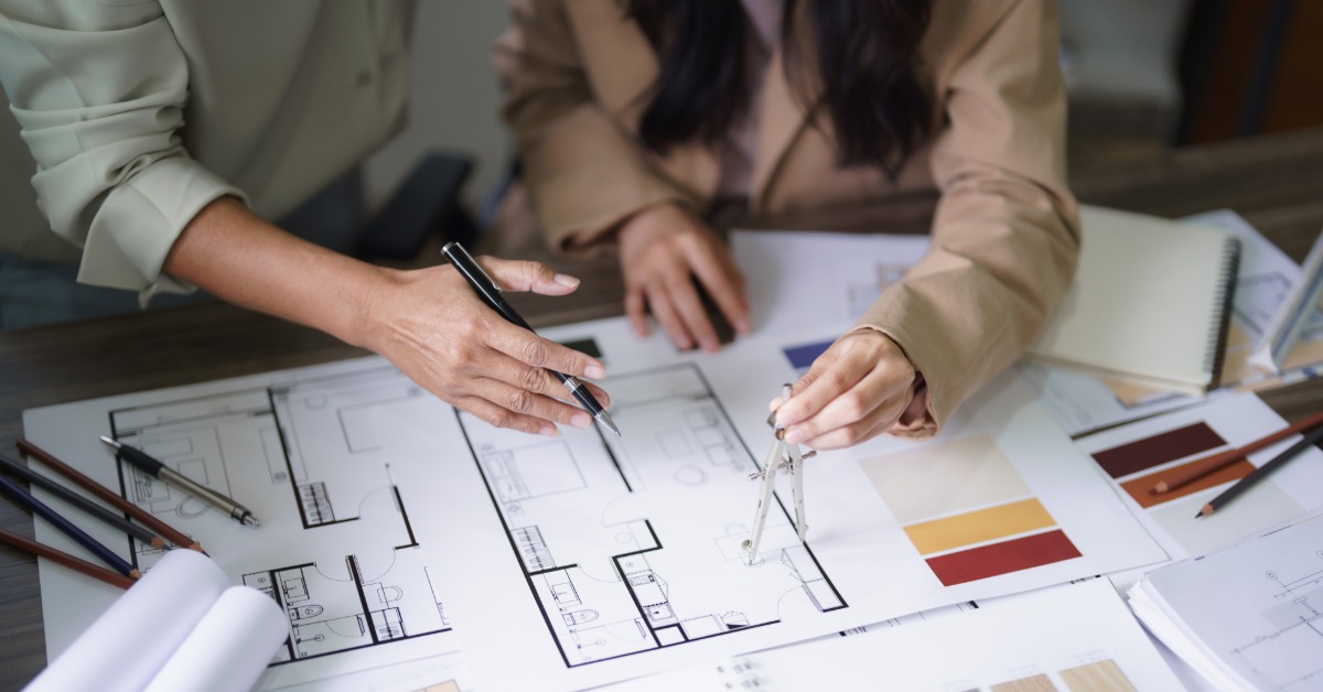 A close-up of a man and a woman sitting at a wooden table looking at drawings and schematics for a home floor plan.