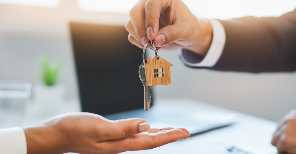 A close-up of a hand holding up a key ring with a key and a miniature home keychain to drop into another hand underneath.
