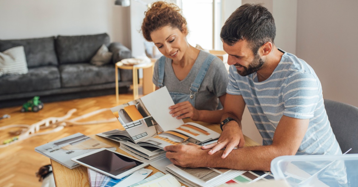 A man and a woman looking at color swatches, a design booklet, and material samples at a table in a home.