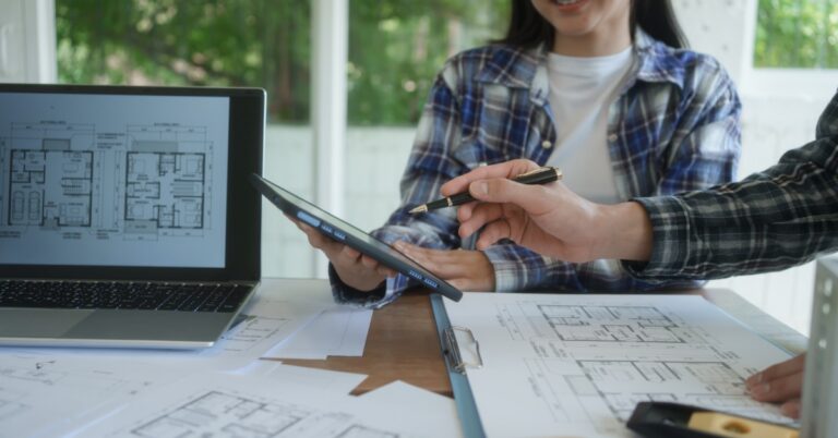 A woman holding an electronic tablet sitting at a table with house floor plans and a laptop while a man points with his pen.