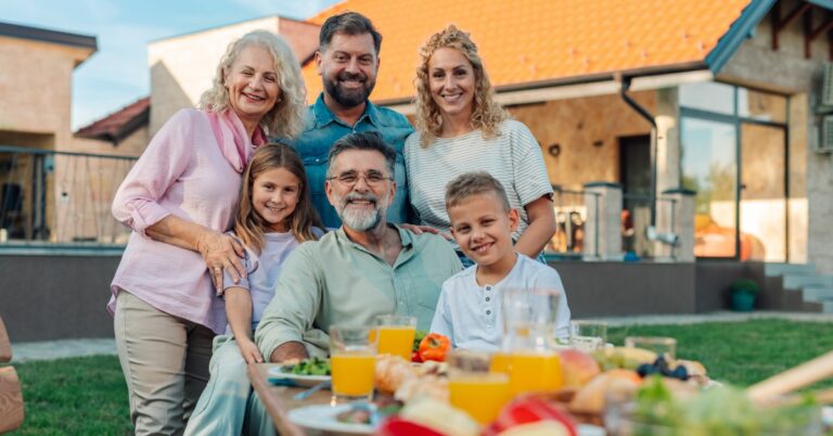 A multi-generational family posing together at the end of a table filled with food and drink in the backyard of a home.