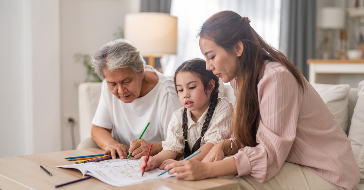 A young girl, an adult woman, and a senior woman are sitting on a couch and coloring in a book together.