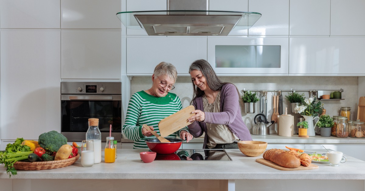 An adult woman and a senior woman cooking together on a kitchen island stove filled with food in a modern kitchen.