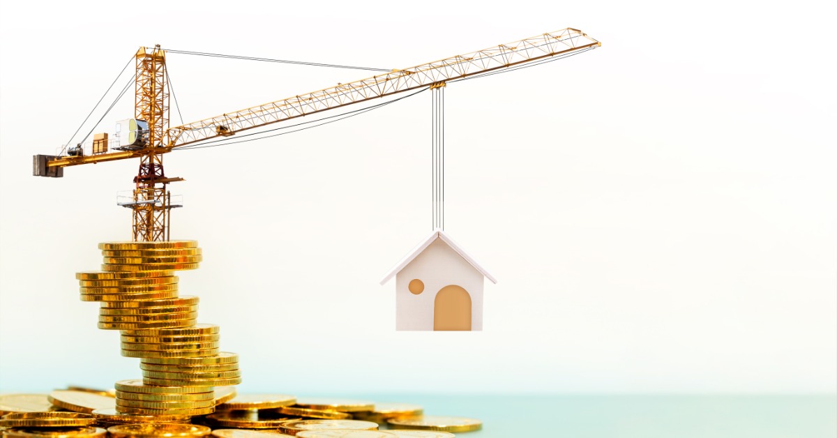 A close-up of a miniature crane on top of a stack of gold coins holding a miniature house model in the air.