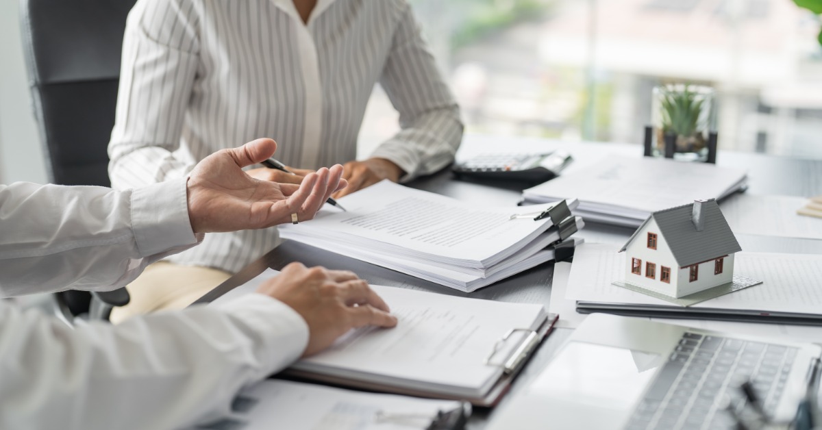 A close-up of a man and a woman at a table filled with clipboards filled with papers, a laptop, and a miniature house.