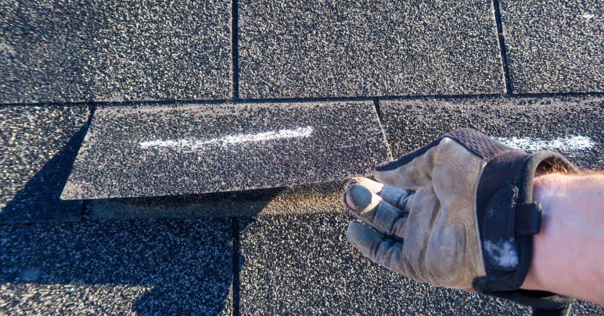 A point-of-view of a person's hand with a glove lifting up a roof shingle marked with white chalk to inspect it.